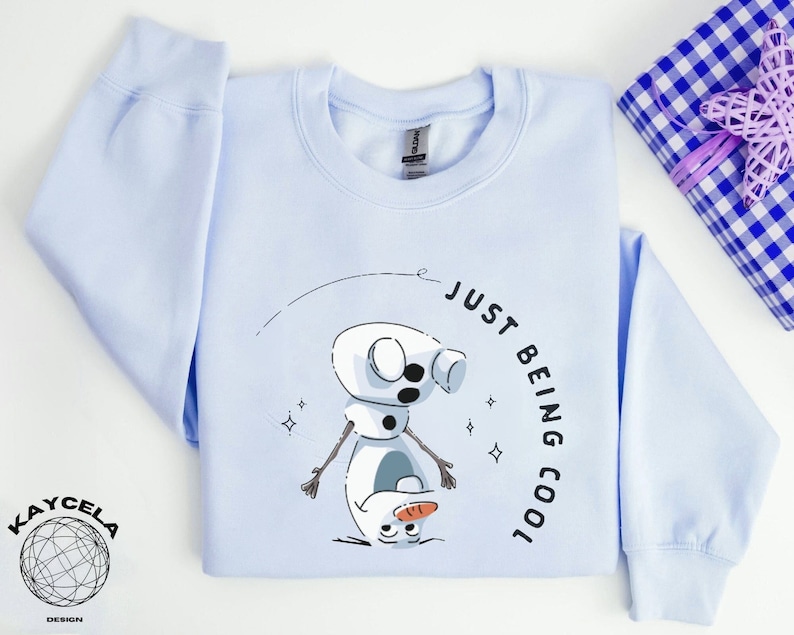 May include: Light blue sweatshirt with a graphic of Olaf from Frozen, the text "Just Being Cool" and a small star design.