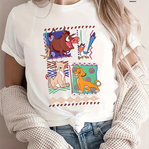 May include: White t-shirt with a colorful graphic print of characters from the movie The Lion King. The characters include Simba, Nala, Pumbaa, and Zazu. The graphic is surrounded by a colorful geometric pattern.