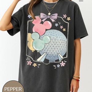 May include: Dark gray t-shirt featuring a graphic of Mickey Mouse balloons, a bow, and a geometric sphere. The design includes pastel pink, blue, and yellow colors. The shirt has the text "PEPPER COMFORT COLORS" at the bottom.
