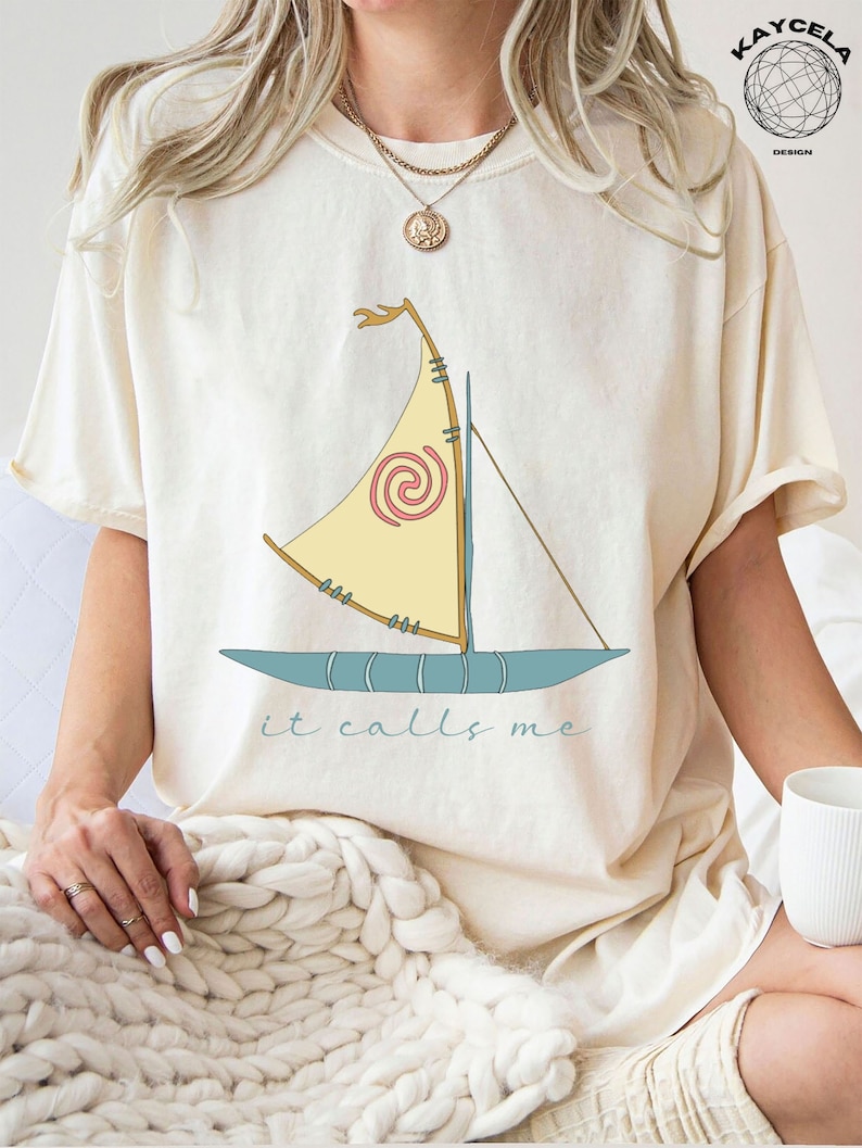 May include: A cream colored t-shirt with a cartoon image of a sailboat with the text "it calls me" printed below the image.