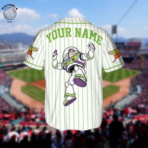Puede incluir: Camiseta de b&eacute;isbol blanca con finas rayas verdes y el texto "YOUR NAME" en la parte superior de la espalda. Presenta un gr&aacute;fico de Buzz Lightyear en la espalda y el logotipo de "Toy Story" en la manga. La camiseta est&aacute; sobre un fondo de campo de b&eacute;isbol.