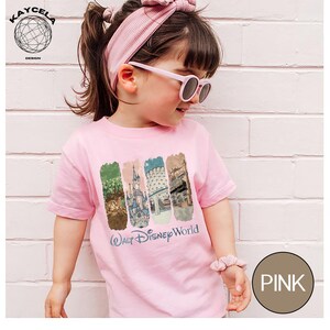 Op de afbeelding: Een roze t-shirt met een "Walt Disney World" afbeelding. De afbeelding toont vier verticale rechthoekige ontwerpen met afbeeldingen van Disney-attracties. Het shirt wordt gedragen door een persoon die een roze zonnebril en een bijpassende haarband draagt.