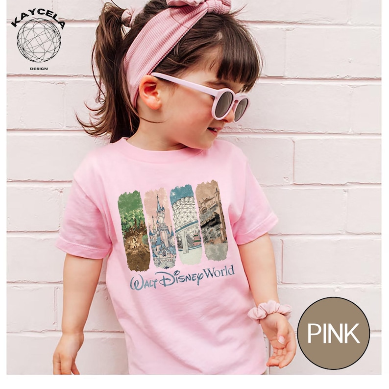 Op de afbeelding: Een roze t-shirt met een "Walt Disney World" afbeelding. De afbeelding toont vier verticale rechthoekige ontwerpen met afbeeldingen van Disney-attracties. Het shirt wordt gedragen door een persoon die een roze zonnebril en een bijpassende haarband draagt.