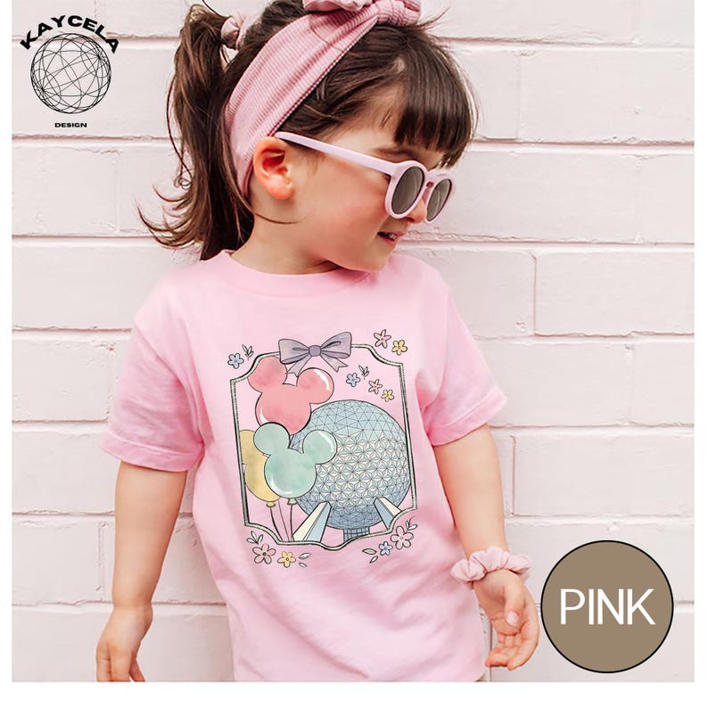 May include: Pink t-shirt featuring a graphic of Mickey Mouse balloons and a geometric sphere. The shirt is adorned with a bow and floral accents. The word "PINK" is in a circle on the bottom right. The shirt is worn by a young child.