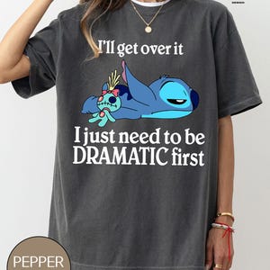 May include: A dark gray t-shirt with a white graphic of Stitch from Lilo & Stitch. The graphic shows Stitch laying down with a doll. The text "I'll get over it I just need to be DRAMATIC first" is printed on the shirt.