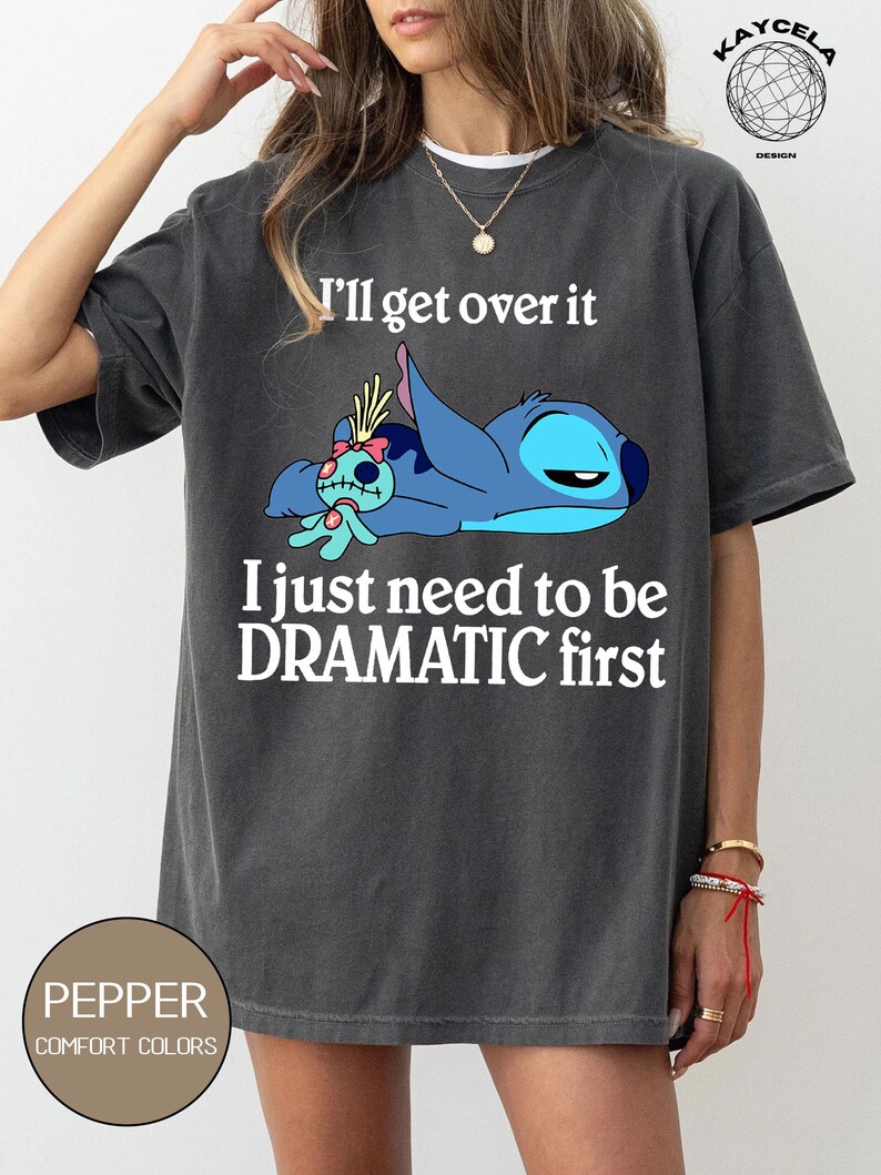 May include: A dark gray t-shirt with a white graphic of Stitch from Lilo & Stitch. The graphic shows Stitch laying down with a doll. The text "I'll get over it I just need to be DRAMATIC first" is printed on the shirt.