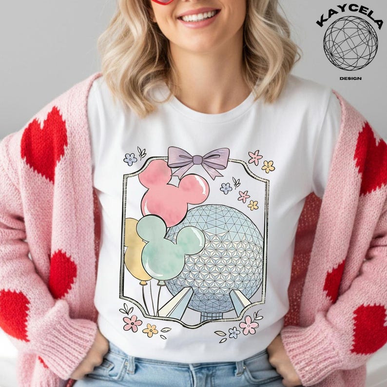 May include: White t-shirt featuring a colorful graphic of Mickey Mouse-shaped balloons, a bow, flowers, and a geometric sphere. The design is framed with a gold border. The shirt is part of a clothing collection.