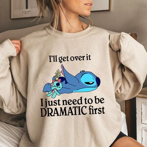 May include: Beige sweatshirt with a blue Stitch character from Lilo & Stitch. The text on the shirt reads "I'll get over it I just need to be DRAMATIC first".