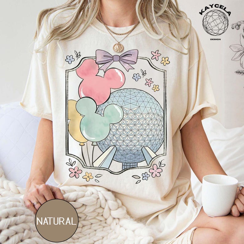 May include: Cream-colored t-shirt featuring a framed graphic with Mickey Mouse balloons in pink, yellow, and mint green, and a geometric sphere. The design includes a purple bow and floral accents. The word "NATURAL" is printed on a brown circle.