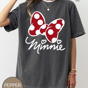 May include: A gray t-shirt with a red and white polka dot bow and the word "Minnie" in white lettering. The shirt has a distressed look and is made of a soft, comfortable material.