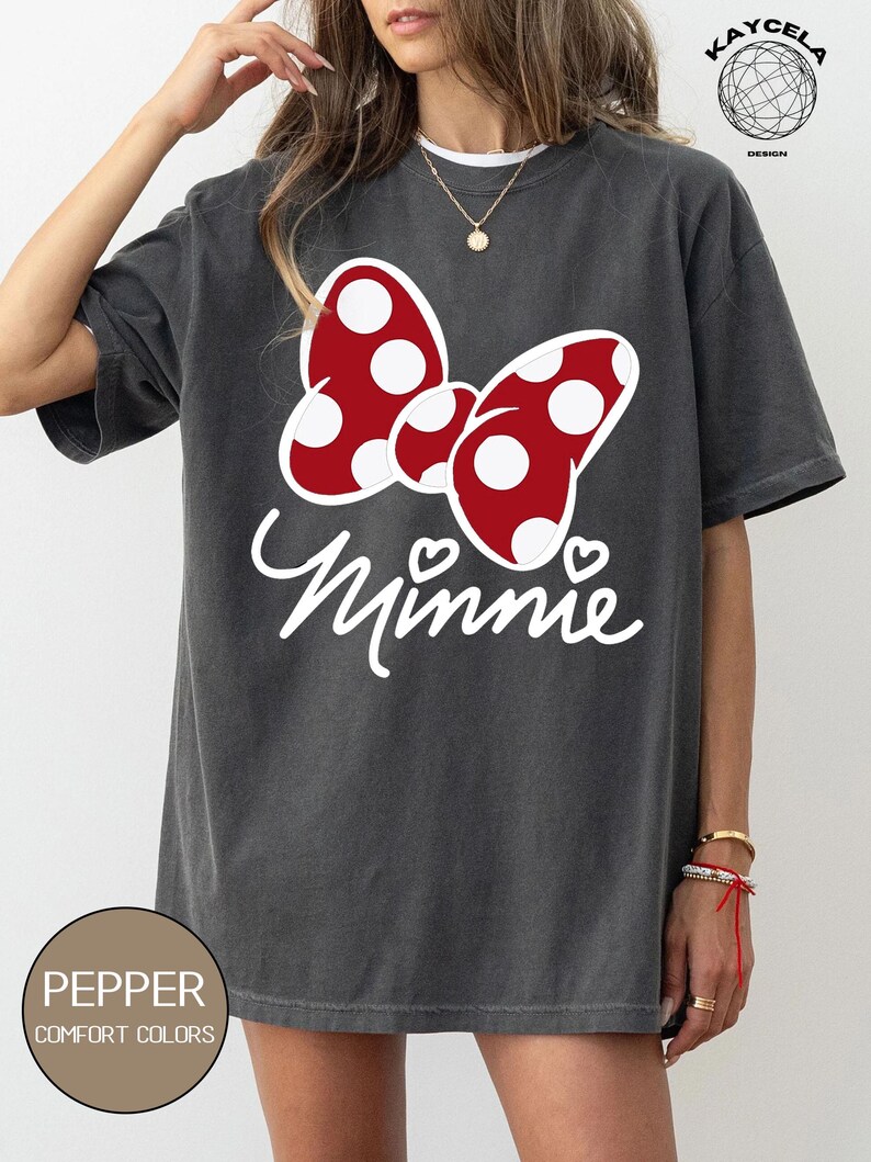 May include: A gray t-shirt with a red and white polka dot bow and the word "Minnie" in white lettering. The shirt has a distressed look and is made of a soft, comfortable material.