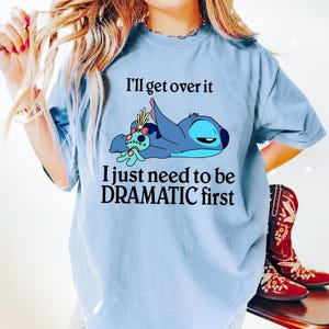 May include: Light blue t-shirt with a cartoon Stitch character from Lilo & Stitch. The text on the shirt reads "I'll get over it I just need to be DRAMATIC first."