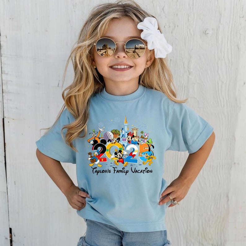 Puede incluir: Una camiseta azul claro con un gr&aacute;fico colorido de personajes de Disney y el texto "2023". La camiseta tambi&eacute;n incluye el texto "Taylor's Family Vacation". La camiseta la lleva una ni&ntilde;a peque&ntilde;a con pelo rubio y gafas de sol.