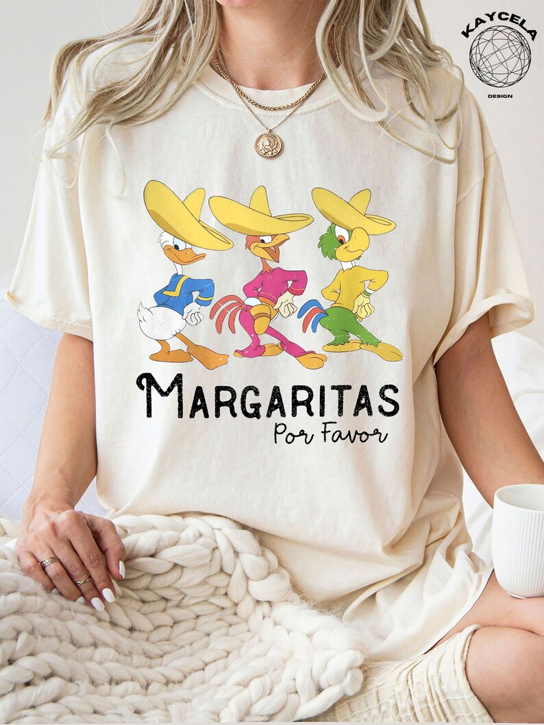 May include: A white t-shirt with a graphic of three cartoon characters wearing sombreros. The characters are a duck, a rooster, and a parrot. The text "Margaritas Por Favor" is printed below the characters.