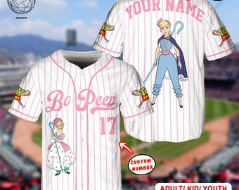 May include: White baseball jersey with pink pinstripes, featuring "Bo Peep" and the number 17 in pink. The jersey has a cartoon image of Bo Peep and the text "YOUR NAME" above the image. The sleeves have a "Toy Story" logo.
