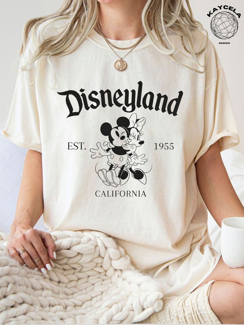 May include: A cream-colored t-shirt with a black graphic of Mickey Mouse and Minnie Mouse kissing. The text "Disneyland" is above the graphic, and "EST. 1955 CALIFORNIA" is below the graphic.