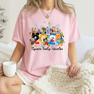 Puede incluir: Camiseta rosa claro con un gr&aacute;fico colorido de personajes de Disney y el texto "2025 Taylor's Family Vacation". El dise&ntilde;o incluye a Mickey Mouse, Donald Duck y otros personajes, con un castillo y fuegos artificiales.