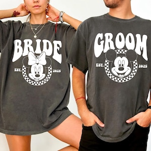 May include: A pair of gray t-shirts with a vintage-style design featuring Mickey Mouse and Minnie Mouse. The shirts say "Groom" and "Bride" with the year "2025" and "EST." printed below the characters.