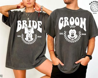 May include: A pair of grey t-shirts with a vintage-style design featuring Mickey Mouse and Minnie Mouse. The shirts say "Groom" and "Bride" with the year "2025" and "EST." printed below the characters.