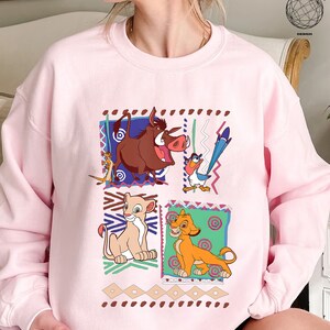 May include: Pink sweatshirt with a graphic print of characters from the Disney movie The Lion King. The characters include Simba, Nala, Pumbaa, and Zazu. The graphic is surrounded by a colorful geometric pattern.