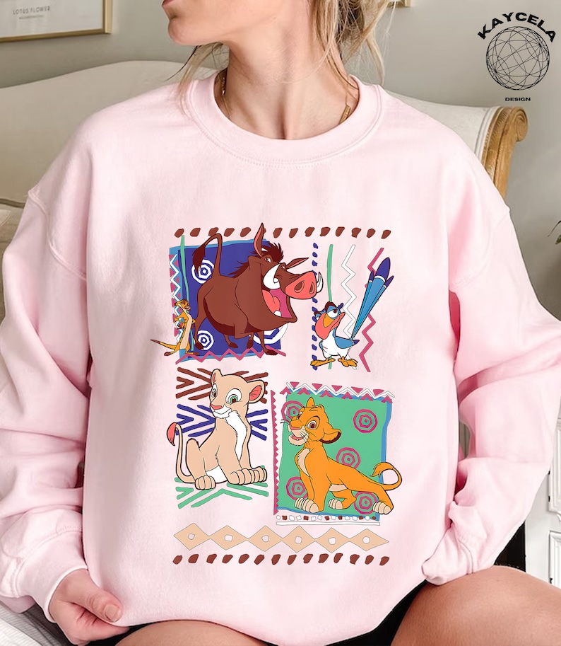 May include: Pink sweatshirt with a graphic print of characters from the Disney movie The Lion King. The characters include Simba, Nala, Pumbaa, and Zazu. The graphic is surrounded by a colorful geometric pattern.
