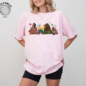 K&ouml;nnte beinhalten: Ein pinkfarbenes T-Shirt mit einer bunten Grafik von Figuren aus dem Film Vaiana. Die Grafik enth&auml;lt die Figuren Maui, Vaiana, Pua und Hei Hei. Die Grafik ist so angeordnet, dass sie das Wort "Vaiana" buchstabiert.