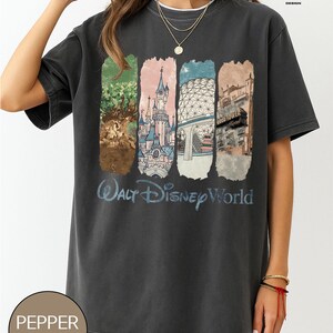 Op de afbeelding: Donkergrijs t-shirt met een kleurrijke afbeelding van Walt Disney World-bezienswaardigheden. Het ontwerp bevat afbeeldingen van een boom, een kasteel, een geodetische bol en een hotel, met "Walt Disney World" in blauw schrift. Het shirt heeft een Pepper Comfort Colors label.