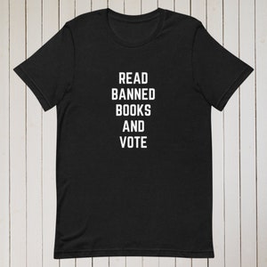 May include: Black t-shirt with white text that reads "READ BANNED BOOKS AND VOTE." The shirt is a crew neck style and appears to be made of a soft material. The background is a light wood grain.