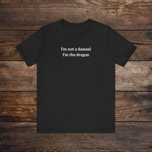 May include: Black t-shirt with the text "I'm not a damsel, I'm the dragon" in white. The shirt is made of a soft material and is suitable for casual wear. The design is centered on the chest.