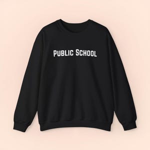 May include: Black crew neck sweatshirt with the words "PUBLIC SCHOOL" printed in white block letters across the chest. The sweatshirt has long sleeves and a ribbed neckline, cuffs, and hem. The garment is made of a soft material.