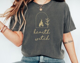 May include: Dark grey t-shirt with a gold-colored graphic of a fireplace, flames, and botanical elements. The words "hearth witch" are written in a script font below the graphic. The shirt is made of a soft, comfortable material.