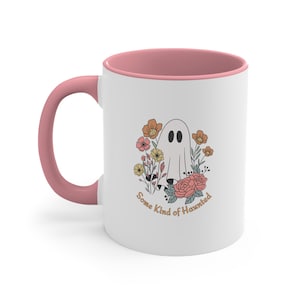 May include: White ceramic mug with a pink interior and handle. The mug features a ghost illustration surrounded by flowers in shades of orange, pink, and yellow. The text "Some Kind of Haunted" is printed on the mug.