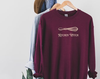 Może przedstawiać: Bordowa bluza z nadrukiem złotego trzepaczki i napisem "Kitchen Witch" poniżej. Bluza wisi na drewnianym wieszaku z innymi ubraniami i kapeluszem w tle.