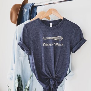 May include: Navy blue t-shirt with a light beige whisk graphic and the words "Kitchen Witch". The shirt is hanging on a wooden hanger with other clothing items and a brown hat in the background.