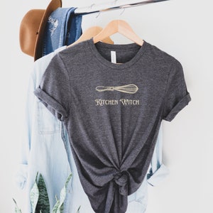 May include: Dark gray t-shirt with a gold whisk graphic and the words "Kitchen Witch". The shirt is hanging on a wooden hanger with a light blue denim shirt and a brown hat in the background.