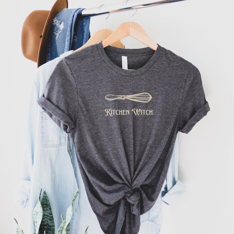 May include: Dark gray t-shirt with a gold whisk graphic and the words "Kitchen Witch". The shirt is hanging on a wooden hanger with a light blue denim shirt and a brown hat in the background.