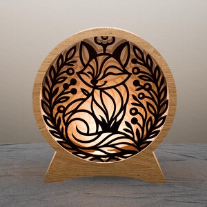 Puede incluir: Lámpara redonda de madera con un diseño de zorro negro rodeado de hojas y bayas. La luz emana a través del diseño del zorro. La lámpara, de unos 20 cm de diámetro, se apoya en una base de madera.