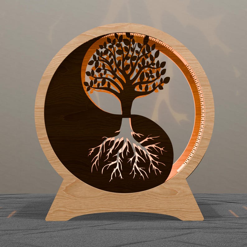 May include: A wooden Yin Yang symbol with a tree design. The tree is cut out of the wood, with the roots in the bottom half and the branches in the top half. The design is illuminated with a warm light.