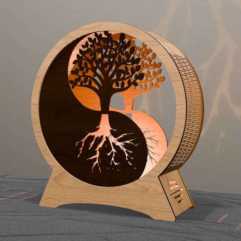 May include: A wooden Yin and Yang lamp featuring a tree design. The lamp has a circular frame with a cutout of a tree, its roots, and branches. The tree is split into two halves, one dark and one light, with an orange glow emanating from within.