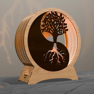 May include: A wooden Yin Yang-shaped lamp featuring a tree design. The lamp has a light brown exterior with a dark brown tree silhouette and roots. The tree's leaves and roots are illuminated, creating a warm glow. The lamp is designed to sit on a flat surface.