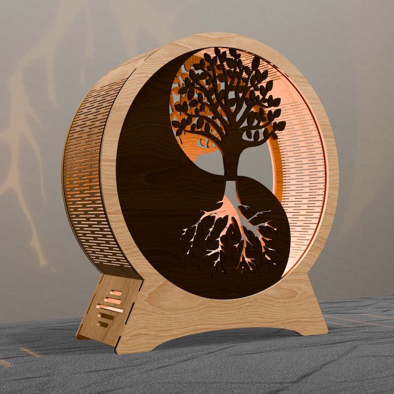 May include: A wooden Yin Yang-shaped lamp featuring a tree design. The lamp has a light brown exterior with a dark brown tree silhouette and roots. The tree's leaves and roots are illuminated, creating a warm glow. The lamp is designed to sit on a flat surface.