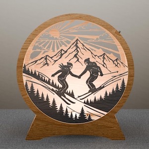 Peut inclure: Lampe décorative ronde en bois avec un motif découpé au laser de deux skieurs se tenant la main sur une pente de montagne enneigée. Le motif comprend un soleil, des montagnes et des pins. La lampe a un socle en bois.