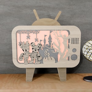 May include: A wooden Halloween-themed light-up TV featuring Mickey and Minnie Mouse dressed as mummies. The design includes a spooky castle, trees, and spiderwebs. The light glows orange, creating a festive ambiance.