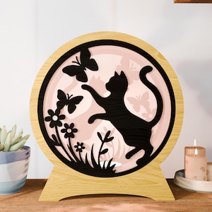 May include: A circular wooden night light with a black cat silhouette chasing butterflies. The inner design has a pink backdrop with floral and butterfly cutouts. The light sits on a wooden base, creating a warm, decorative accent.