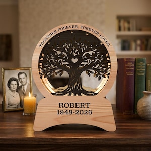Pode incluir: Uma placa memorial circular de madeira com uma silhueta de árvore cortada a laser e um coração no centro. O texto TOGETHER FOREVER, FOREVER LOVED. está no topo, e ROBERT 1948-2026 na base. O design inclui iluminação interna quente.
