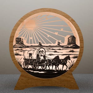 May include: A wooden, circular lamp featuring a laser-cut design of a covered wagon pulled by horses, set against a desert landscape with a sunburst and mesas. The lamp has a wooden base and frame, creating a warm, rustic aesthetic.