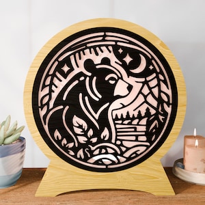 May include: A round wooden lamp with a black bear silhouette design. The bear is surrounded by a forest scene with a moon and stars. The lamp has a light brown wooden base and a circular frame.