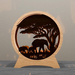 May include: A wooden, circular lamp featuring a laser-cut silhouette of elephants under a tree on the savanna. The lamp has a warm glow, and the frame is made of light-coloured wood.