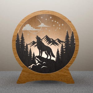 May include: Wooden, round-shaped night light featuring a howling wolf silhouette against a mountain and forest backdrop. The design includes a moon, stars, and clouds. The light is encased in a wooden frame with a stand.