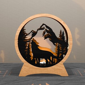 May include: A wooden, circular lamp featuring a layered design of a howling wolf, mountains, and trees. The wolf is silhouetted against a warm, glowing background. The lamp has a natural wood frame and a stand.